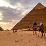 Three Tourists Are Riding Camel And Moving To The Pyramid
