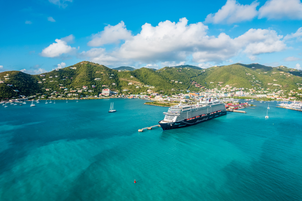 Das Bild zeigt ein großes Kreuzfahrtschiff der „Mein Schiff“-Flotte, das in einem türkisblauen Hafen vor Anker liegt. Im Hintergrund sieht man eine grüne, hügelige Insel mit vielen kleinen weißen Häusern, verteilt zwischen Palmen und Bäumen. Am Ufer befinden sich weitere Boote und Yachten. Der Himmel ist blau mit ein paar weißen Wolken – die Szene wirkt tropisch, sonnig und einladend.
